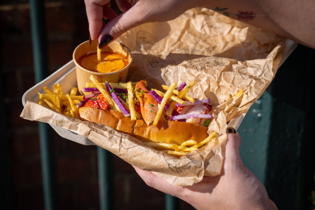 Lobster roll topped with red onion and fries, served with a pot of dipping sauce.
