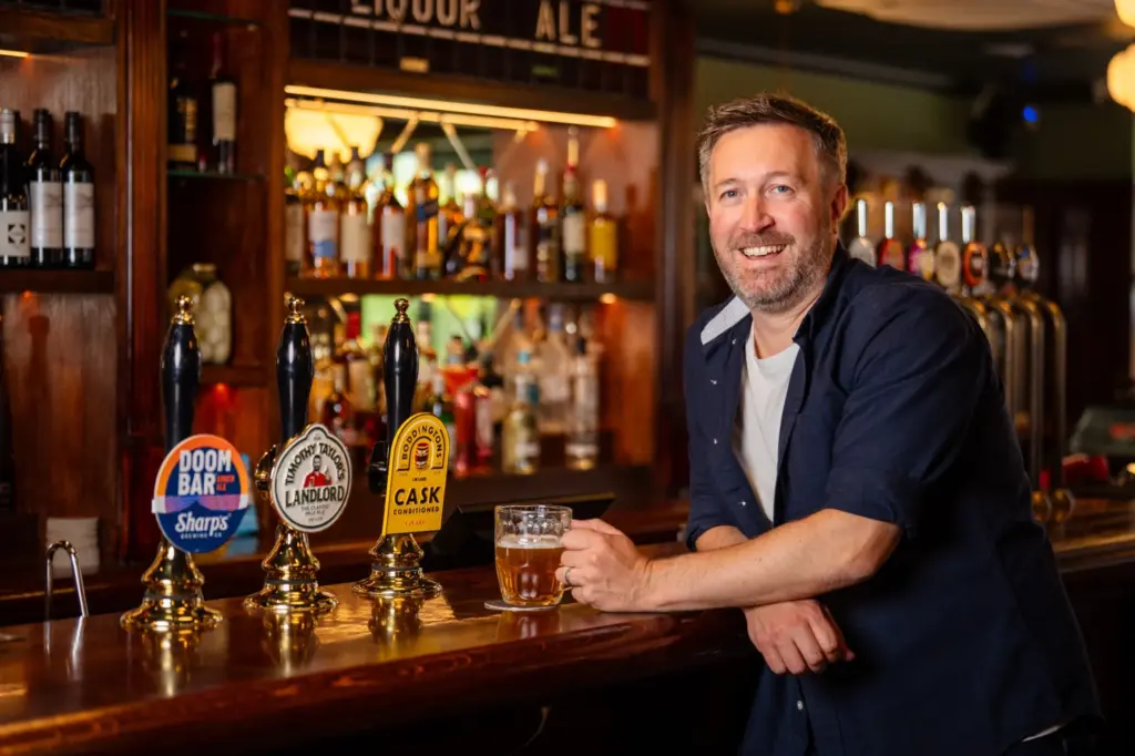Man smiling at the bar with a pint of beer and beer pumps in front of him.